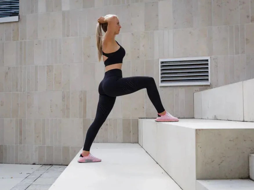 Blonde sportliche Frau posiert in schwarzer Sport Leggings und schwarzem Sports Bra von raise Sportswear auf großer Treppenstufe