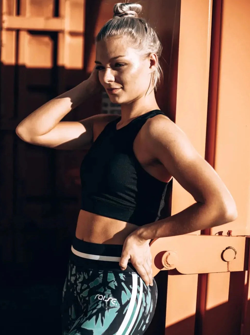 Blonde sportliche Frau in grün schwarzer Sport Leggings und schwarzem Sport BH von raise Sportswear posiert vor orangem Frachtcontainer