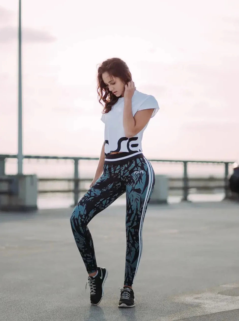 Brünette schlanke Frau posiert in grün schwarzer Sport Leggings und weißem Crop Top von raise Sportswear posiert auf Parkdeck