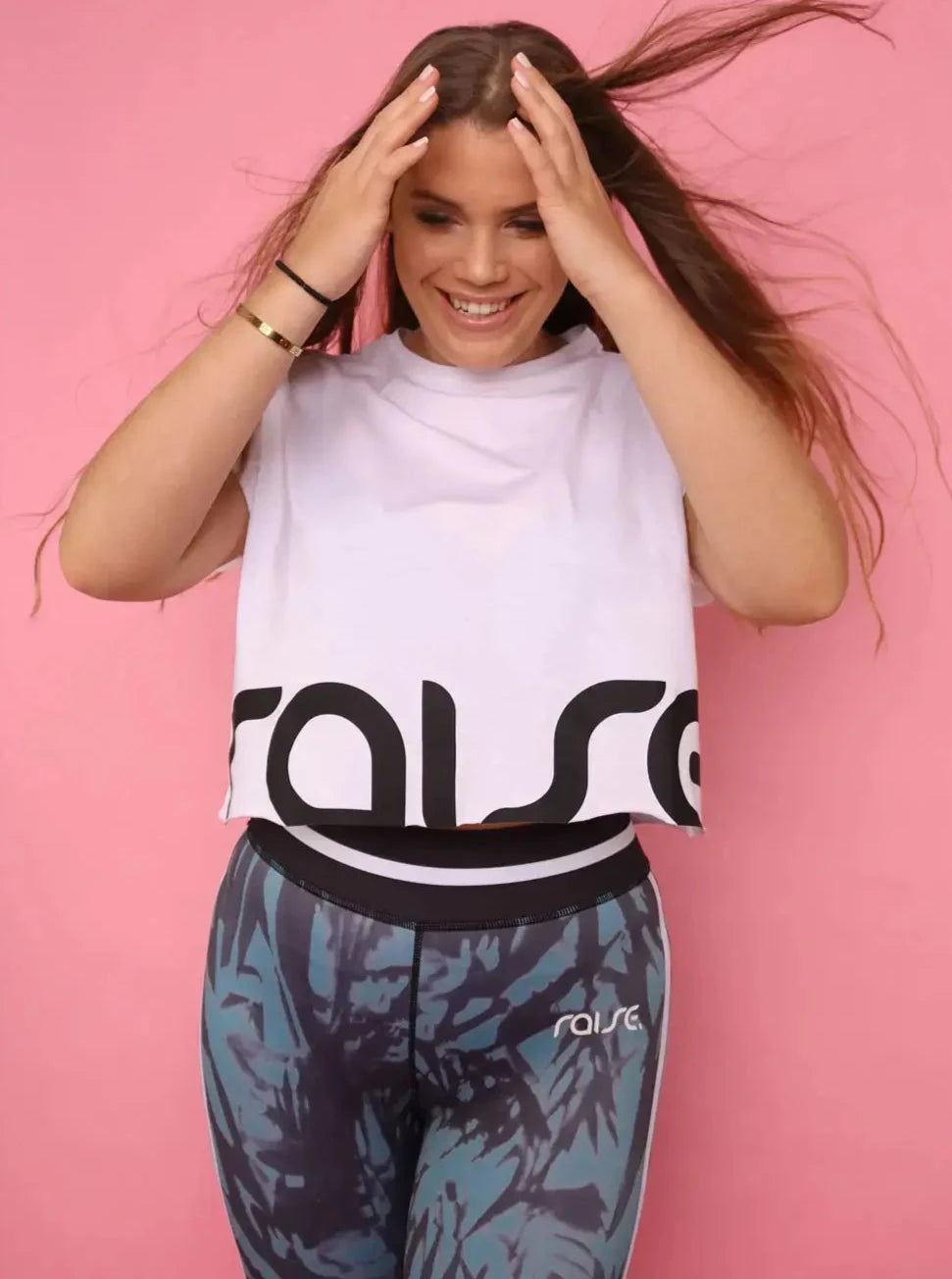 Dunkelblonde kurvige Frau posiert in schwarz grüner Sport Leggings und weißem Crop Top posiert vor pinkem Hintergrund