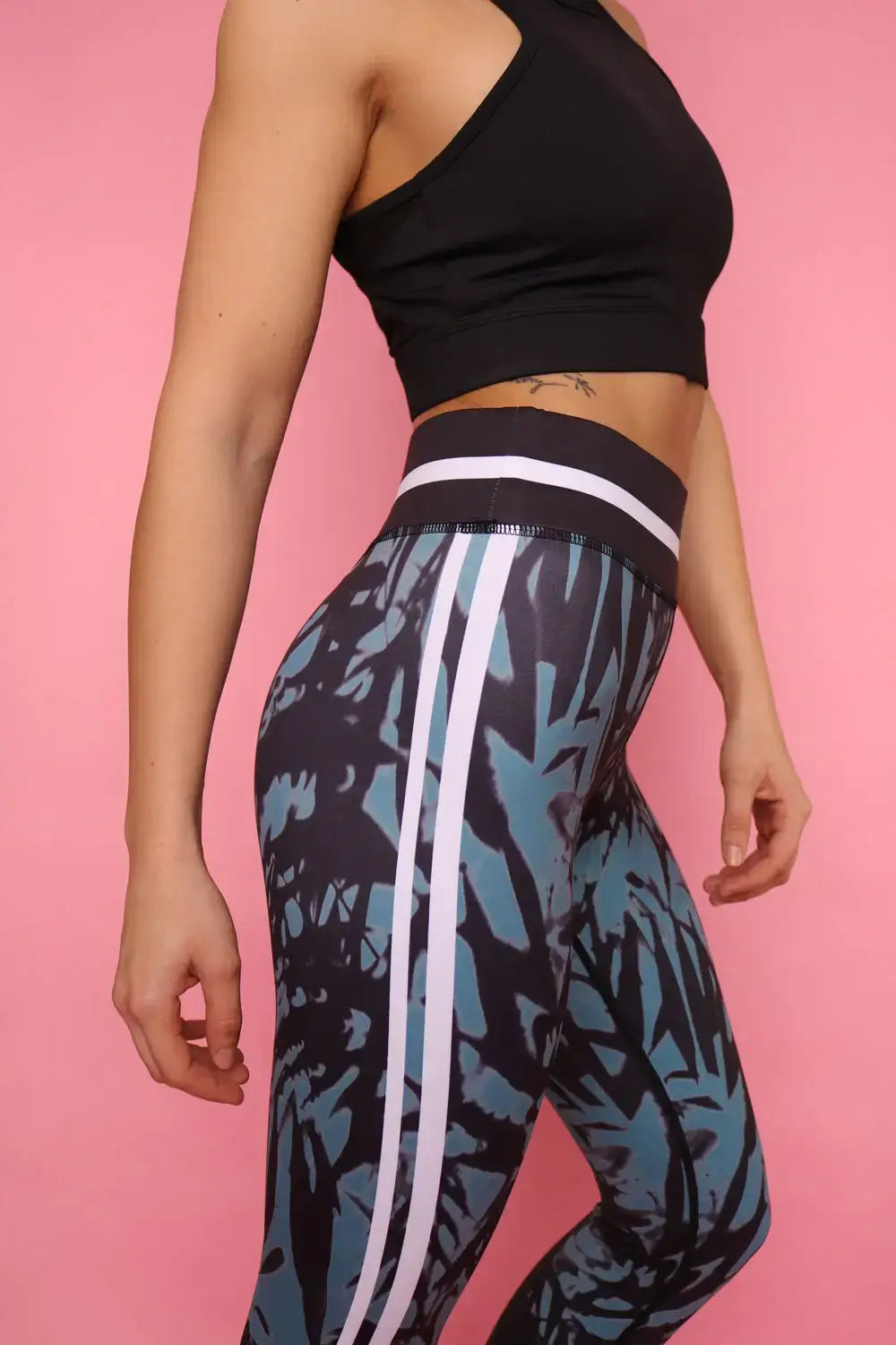 Sportliche Frau in schwarz grüner Sport Leggings und schwarzem Sports Bra von raise Sportswear steht seitlich vor pinken Wand