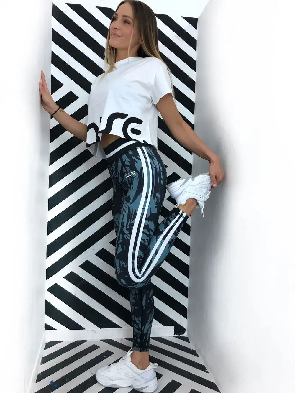 Blonde schlanke Frau in grün schwarzer Sport Leggings und weißem Crop Top von raise Sportswear posiert vor schwarz weiß gestreiften Wand