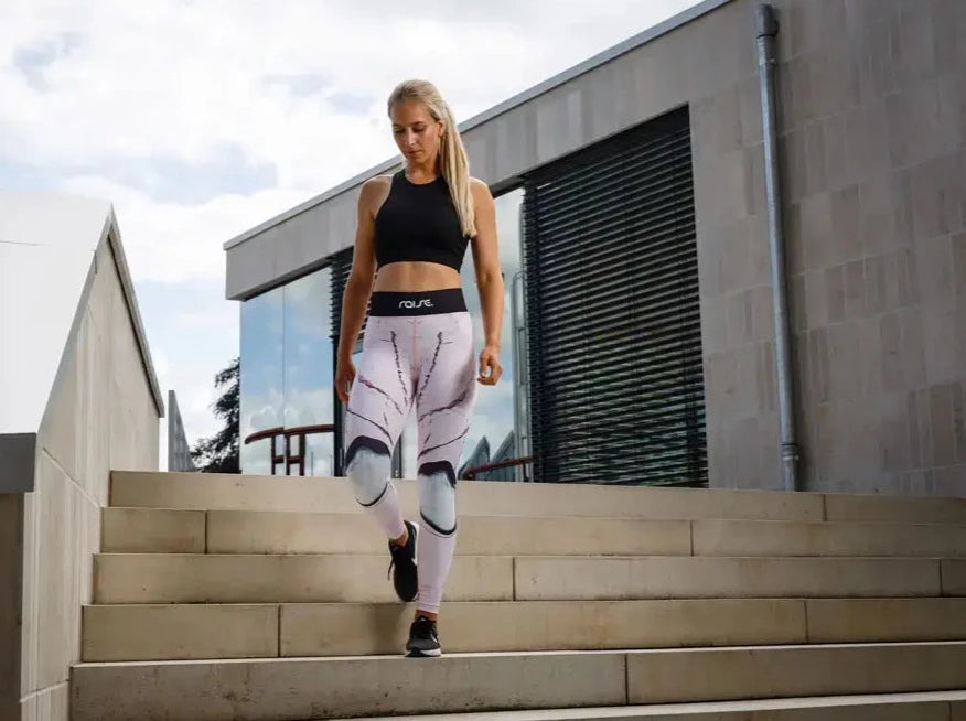 Blonde sportliche Frau in rosa Sport Leggings und schwarzem Sport BH von raise Sportswear geht draußen Treppenstufen runter