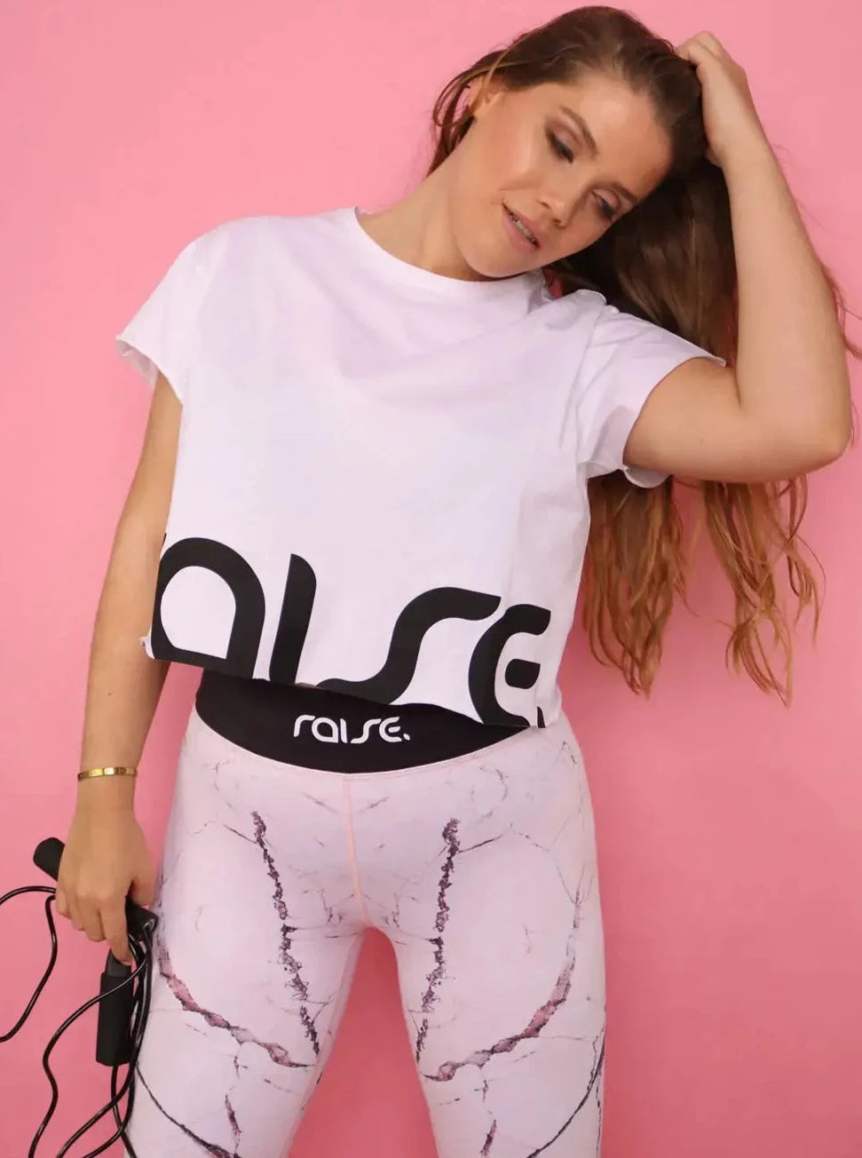 Kurvige dunkelblonde Frau in rosa Sport Leggings und weißem Crop Top von raise Sportswear posiert vor pinken Hintergrund