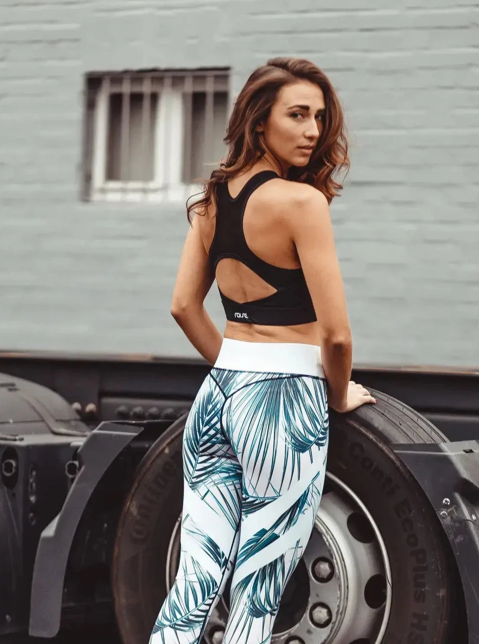 Brünette schlanke Frau in rosa Sport Leggings mit Palmenmuster und schwarzem Sports Bra von raise Sportswear posiert an LKW Reifen