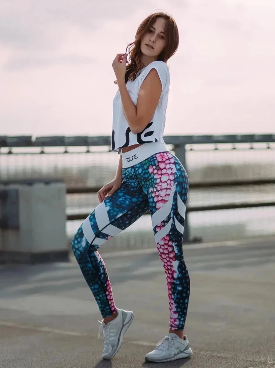 Brünette schlanke Frau in bunter Sport Leggings und weißem Crop Top von raise Sportswear posiert auf Parkdeck