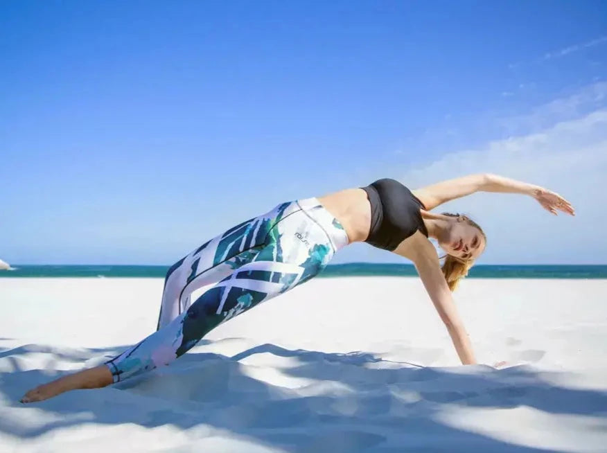 Sportliche Frau in bunter Sport Leggings und schwarzem Sport BH von raise Sportswear macht Yoga am Strand