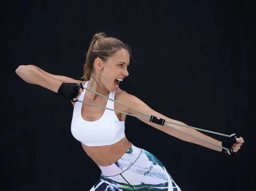 Frau in bunter Sport Leggings und weißem Sports Bra von raise Sportswear dehnt Fitnessband vor schwarzem Hintergrund