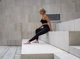 Blonde sportliche in schwarzer Sport Leggings und Sports Bra von raise Sportswear sitzt auf großer Treppenstufe draußen