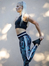 Blonde sportliche Frau posiert in grün schwarzer Sport Leggings und schwarzem Sport BH von raise Sportswear dehnt sich an Wand