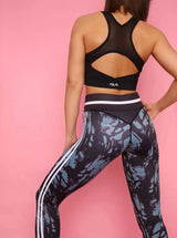 Brünette sportliche Frau in schwarz grüner Sport Leggings und Sport BH von raise Sportswear posiert von hinten vor pinker Wand