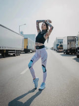 Schlanke tätowierte Frau in rosa Sport Leggings und schwarzem Sports Bra posiert auf Straße mit LKWs