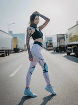 Schlanke tätowierte Frau in rosa Sport Leggings und schwarzem Sports Bra posiert auf Straße mit LKWs