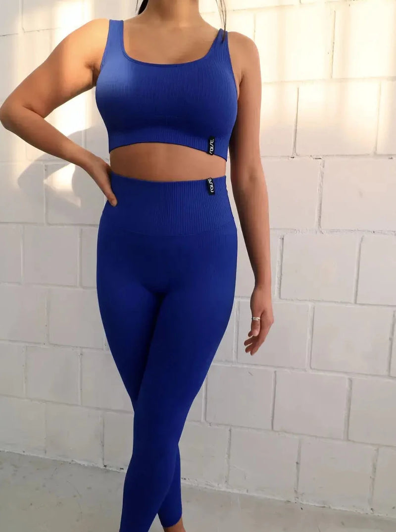Brünette Frau in blauer Seamless Sport Leggings und blauem Sports Bra von raise Sportswear posiert vor weißer Wand